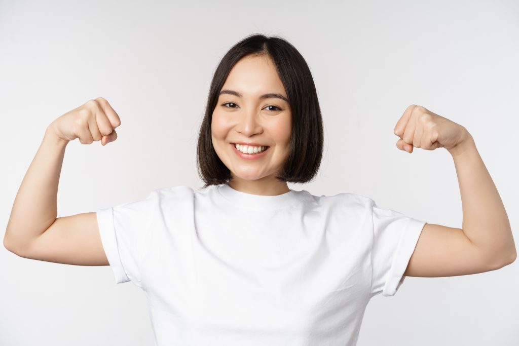 Smiling asian woman showing flexing biceps, muscles strong arms gesture, standing in white tshirt. Strong immune system