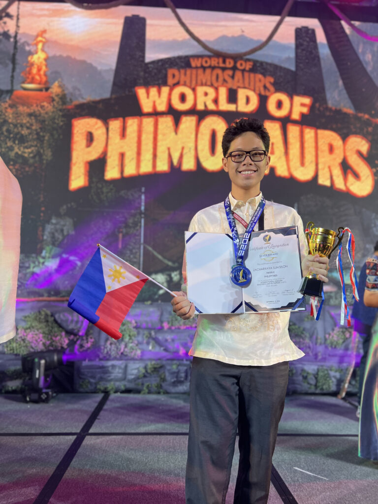 Zacharraya Sun Sison holding the Philippine flag, a trophy, and his medal from IEMO 2026